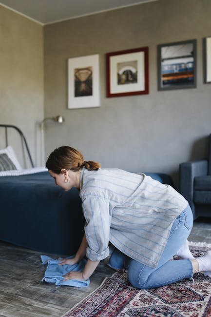 Close-up of a black vacuum cleaner head being used on a patterned, cream-colored carpet in a residential room, with a person wearing white sneakers visible in the background. The carpet features a flo