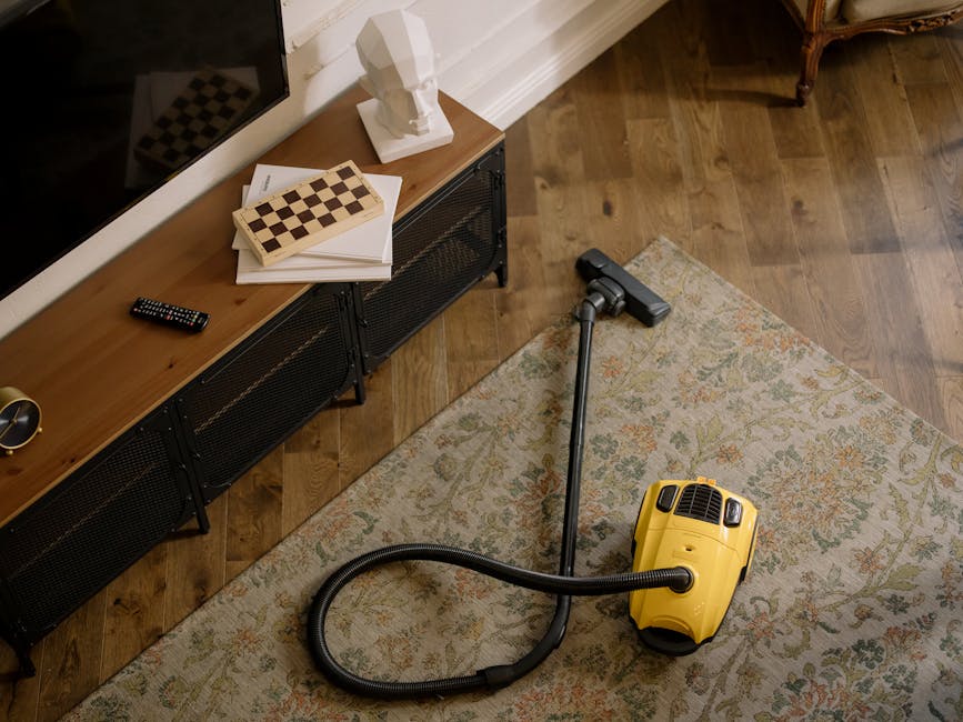 A professional cleaning technician dressed in white protective coveralls, face mask, and blue gloves using a steam or fogging device to sanitise a plush beige carpet in a well-lit living room. The roo