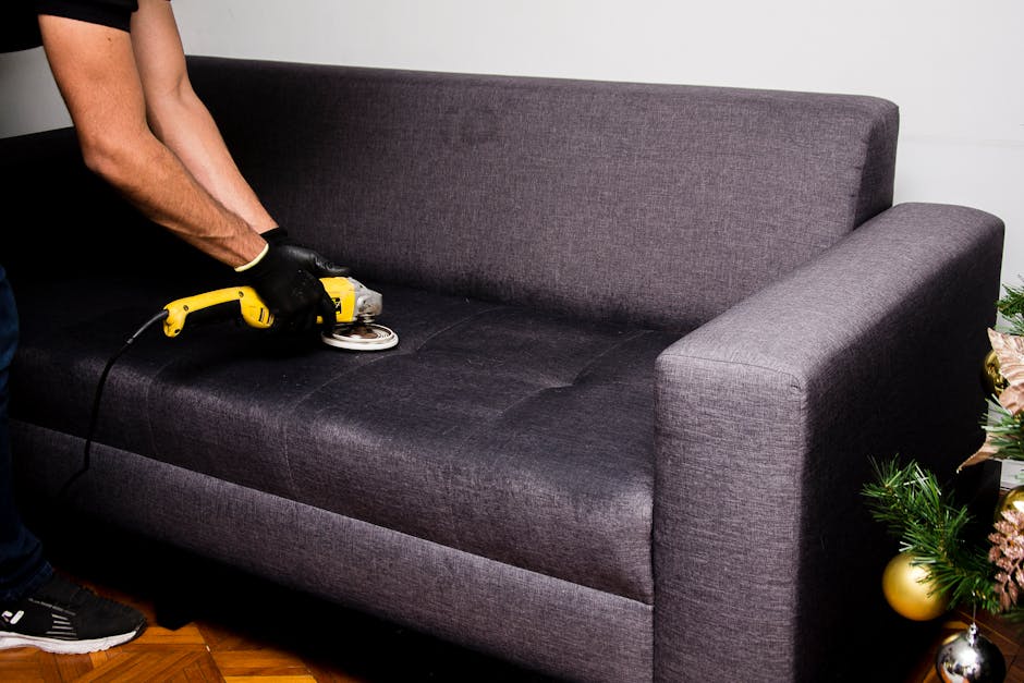 A professional cleaning specialist from Dulwich Carpet Cleaning, dressed in white protective clothing, gloves, and a face mask, is engaged in surface cleaning of a black leather sofa in a well-lit liv