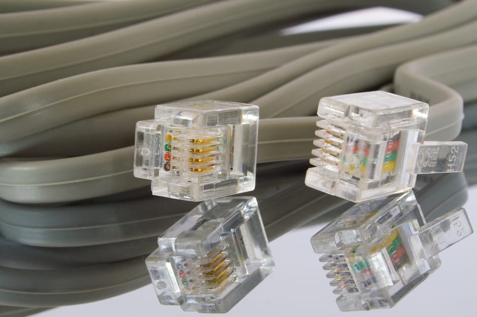 Close-up view of a neatly coiled light gray Ethernet cable resting on a reflective surface, with four transparent RJ45 connectors placed on top. The connectors reveal gold-plated contact pins and colo
